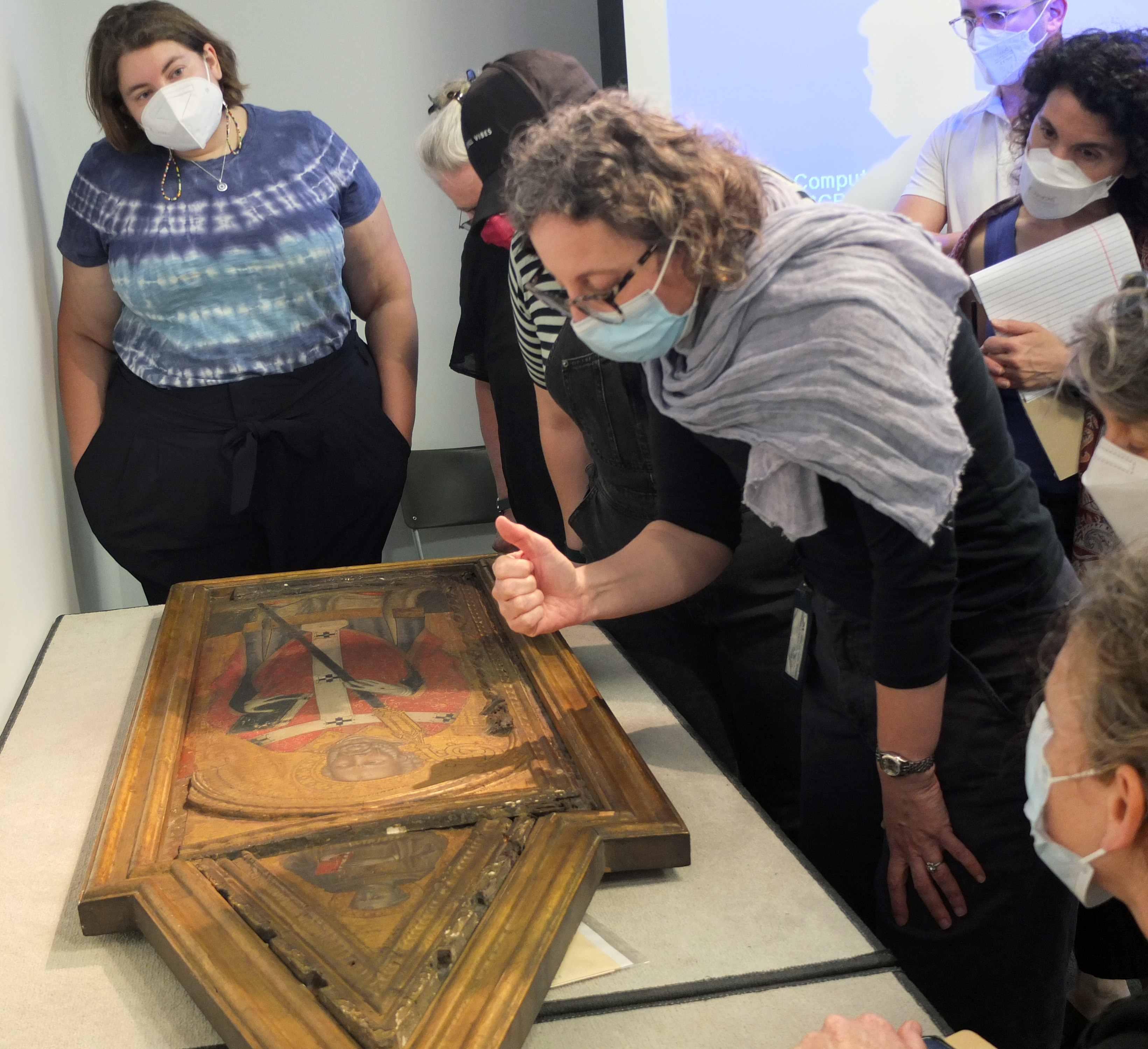 Interior view of a group of standing STITAH participants and leaders examining a gold-framed Medieval painting on a table in YUAG.